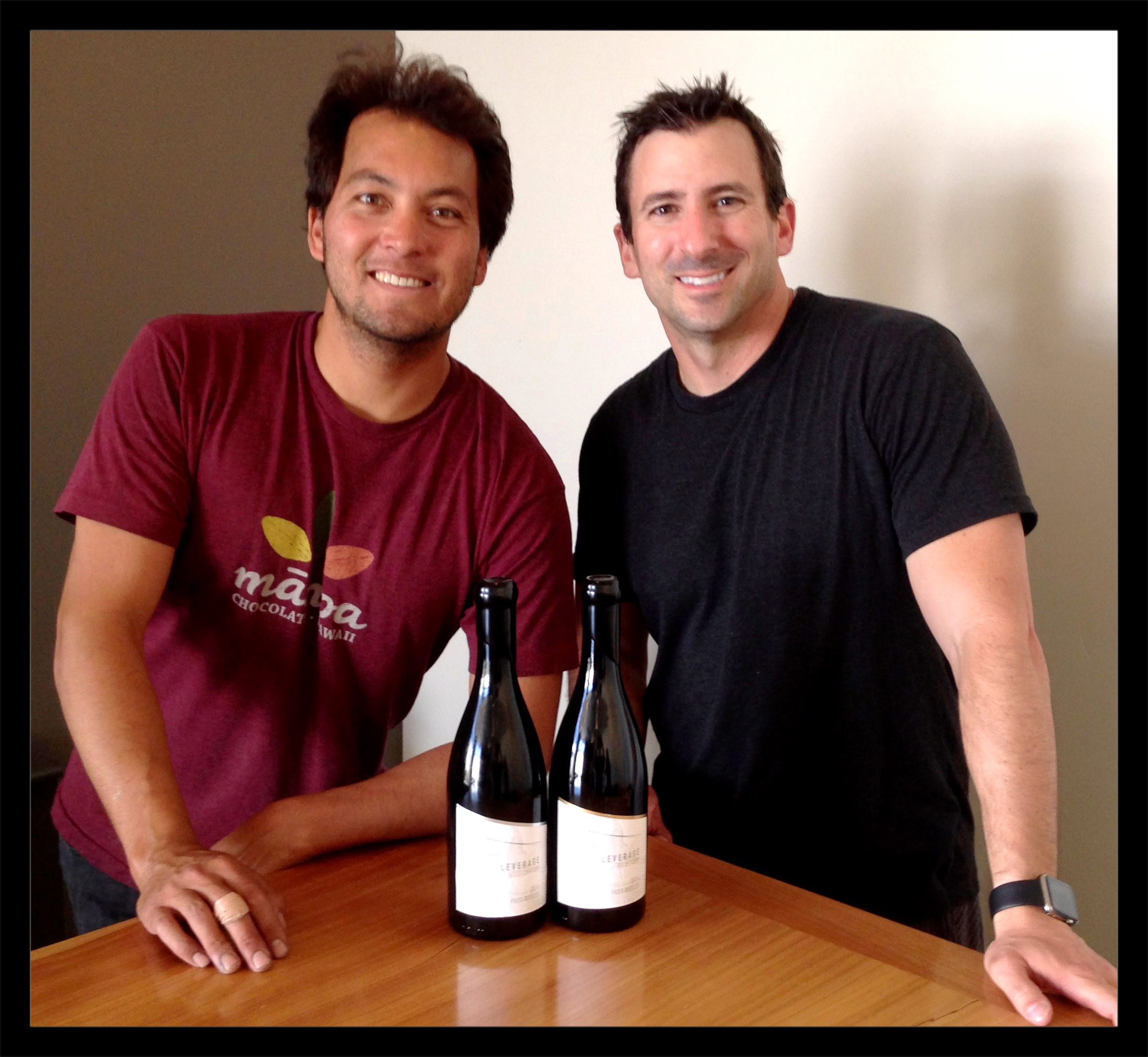 Two smiling men standing behind a table with two wine bottles, celebrating teamwork.